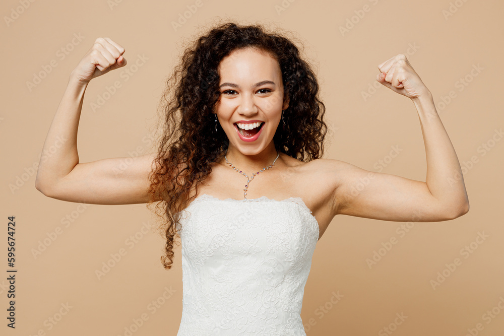 Happy young woman bride wear white wedding dress posing showing muscles on hand demonstrate strength power isolated on plain pastel light beige background studio. Ceremony celebration party concept. Happy young woman bride wear white wedding dress posing showing muscles on hand demonstrate strength power isolated on plain pastel light beige background studio. Ceremony celebration party concept.