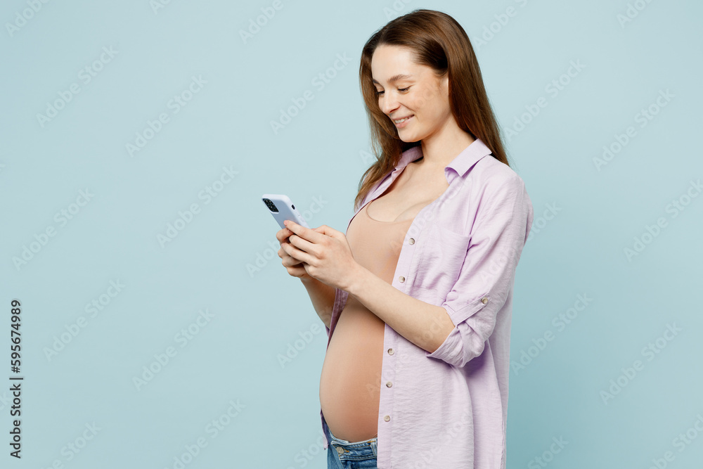 Side view young fun pregnant future mom woman with belly tummy with baby wear casual clothes hold in hand use mobile cell phone isolated on plain pastel blue background. Maternity pregnancy concept. Side view young fun pregnant future mom woman with belly tummy with baby wear casual clothes hold in hand use mobile cell phone isolated on plain pastel blue background. Maternity pregnancy concept.