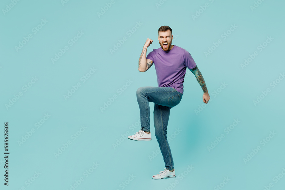 Full body side view happy fun young man he wearing purple t-shirt do winner gesture raise up leg isolated on plain pastel light blue cyan background studio portrait. Tattoo translates life is fight.