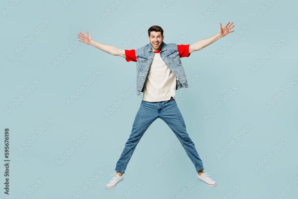 Full body excited happy cool young man he wearing denim vest red t-shirt casual clothes jump high with outstretched hands isolated on plain pastel light blue cyan background studio. Lifestyle concept.