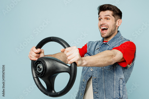 Side view young happy man wear denim vest red t-shirt casual clothes hold steering wheel driving car look camera isolated on plain pastel light blue cyan background studio portrait. Lifestyle concept.
