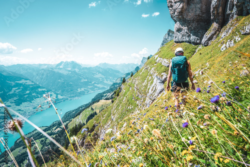 Back view of mountaineer hiking on flowery trail next to steep rock wall with panoramic view on Walensee and the swiss alps in the background. Schnürliweg, Walensee, St. Gallen, Switzerland, Europe.