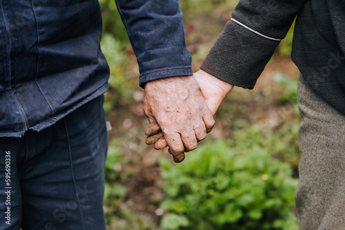 Elderly man and woman gardeners hold hands while standing outdoors in a garden in nature. Closeup photo, eternal love concept.
