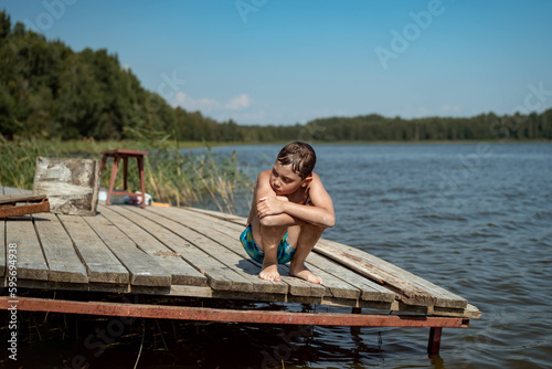 cute caucasian boy sitting on wooden pier sad and cold after swimming in lake