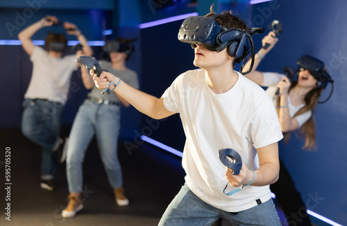 Cheerful emotional teenager in VR headset fully immersed in game, actively engaged in virtual reality world, manipulating objects or shooting targets with joysticks in hands..