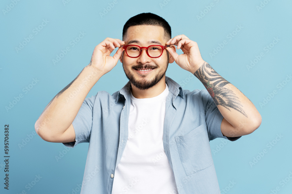 Portrait of handsome smiling asian man with stylish tattoo isolated on blue background. Happy Korean hipster wearing red eyeglasses looking camera. Vision concept