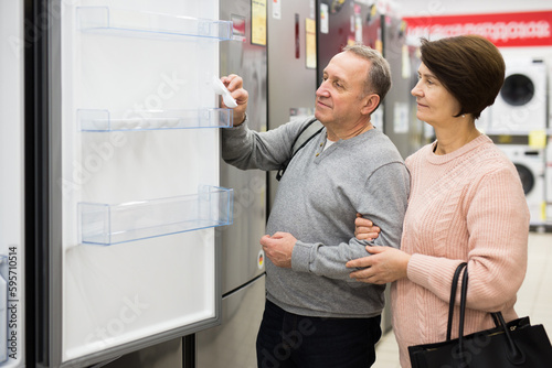 European spouses of mature age, who have come to the electronics and household appliances store for shopping, choose a ..refrigerator, examining it