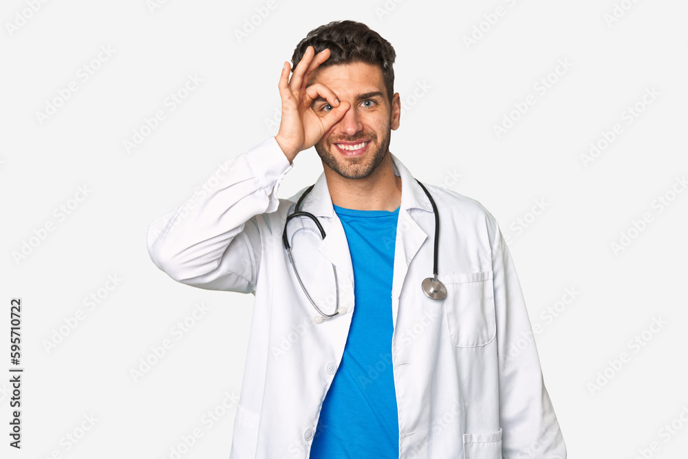 Young doctor man excited keeping ok gesture on eye. Young doctor man excited keeping ok gesture on eye.