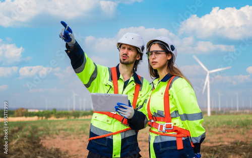 Two professional maintenance engineers in uniform and safety workwear using tablet to checklist repair wind turbines on windmill farm site.
