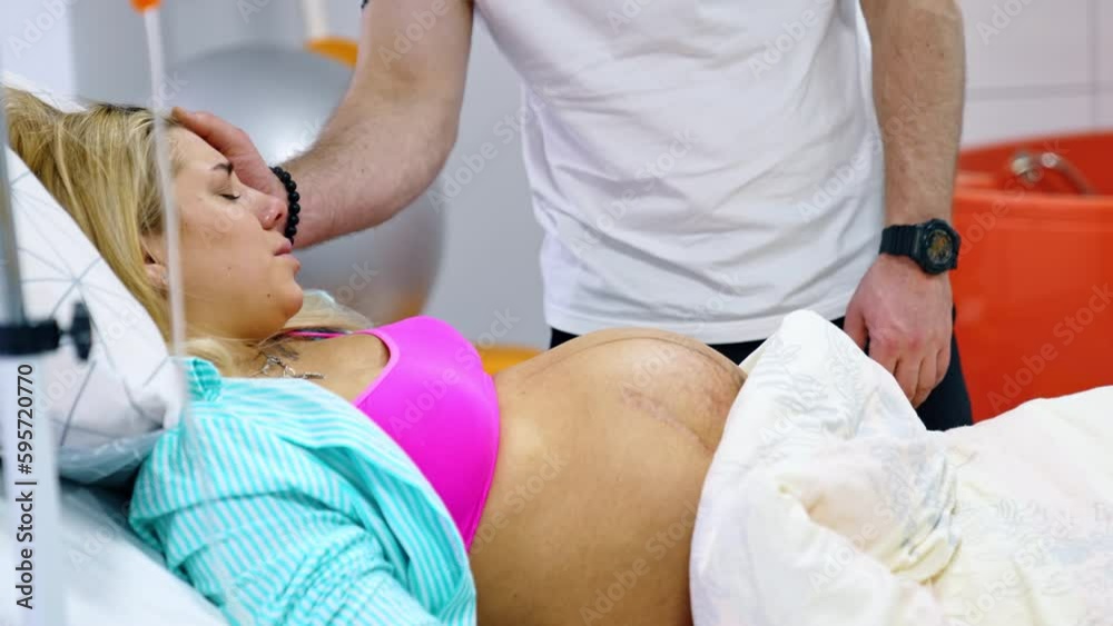 Pregnant Caucasian woman suffers from contractions. Lady lies in bed and her husband stands behind stroking her head. Pregnant Caucasian woman suffers from contractions. Lady lies in bed and her husband stands behind stroking her head.