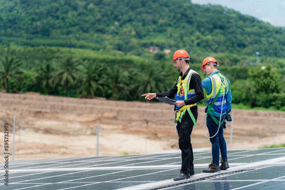 Two young engineer and inspector wearing safety vest and hard helmet inspecting solar panel by kneeling and filling holes and fixing damage using a drill machine while holding a clipboard