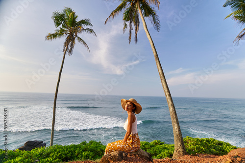 Young african female model posing in colorful clothes at scenic tropical location by ocean between palms at sunrise. Black woman sitting against exotic scenery with view of sea and palm trees at dawn.