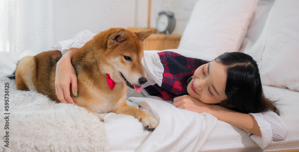 Beautiful asian woman lying on a white bed in the bedroom while embracing a dog with a smile on her face.