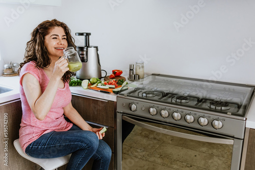 Latin woman drinking green juice and processing ingredients in the kitchen at home in Mexico, Hispanic people