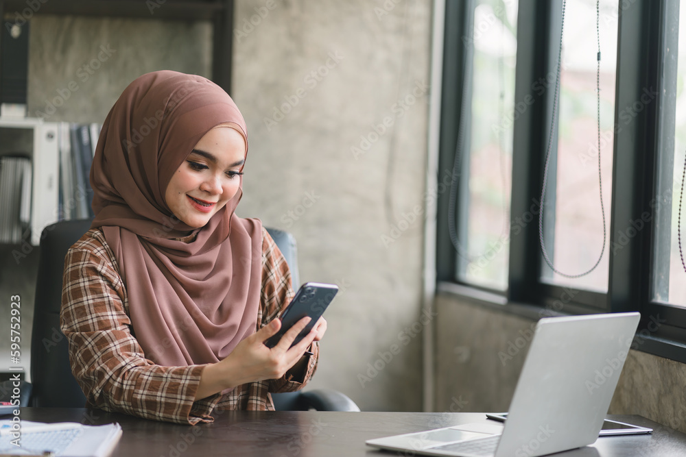 Beautiful muslim business woman wearing a hijab is diligently working at her smartphone and computer, displaying professionalism and commitment to her job. Beautiful muslim business woman wearing a hijab is diligently working at her smartphone and computer, displaying professionalism and commitment to her job.