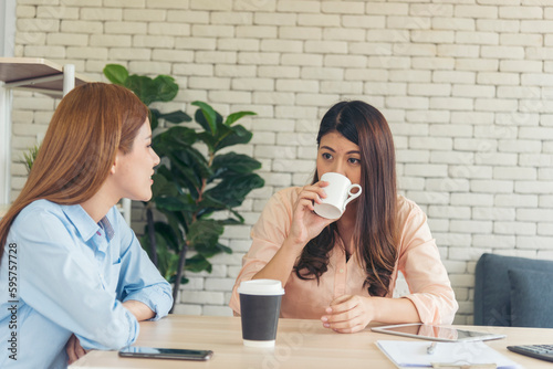 Two Women colleagues drinking black coffee cup at office desk together. Asian woman friendstalking smiling with friendship happy time. Teamwork happy moment lifestyle. International Woman day concept photo