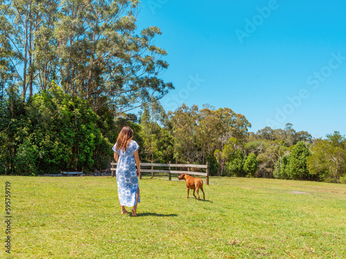 Middle age woman bare feet long brown hair wearing long blue dress back view walking red rhodesian ridgeback hound dog on green field in bush  Pomona country side Sunshine Coast Australia