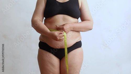 Unrecognizable obese woman wearing black underwear, measuring flabby waist with yellow tape showing result on white background. Weight control, plastic surgery, liposuction, adiposity, body positive. 