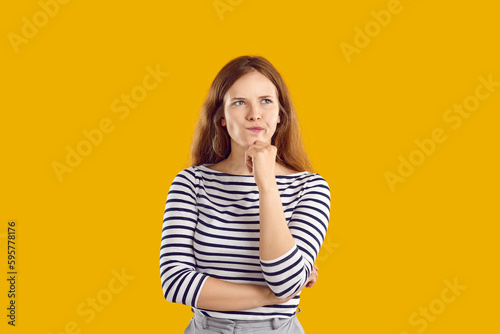 Portrait of serious confused undecided young woman full of doubt holding hand on chin and thinking, trying to make hard choice decision. Girl with wavy hair in striped sweatshirt on yellow background. photo