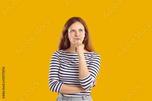Portrait of serious confused undecided young woman full of doubt holding hand on chin and thinking, trying to make hard choice decision. Girl with wavy hair in striped sweatshirt on yellow background.