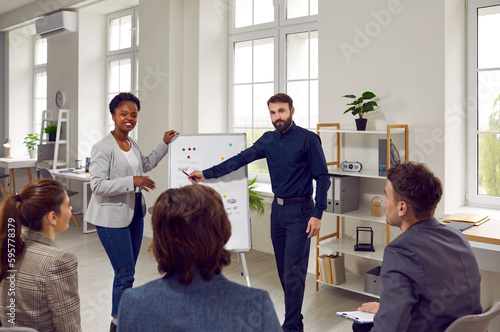 Young professional team employees of progressive company oversees presentation of colleagues from marketing or finance department stand in spacious office next to flipchart. Multiracial people