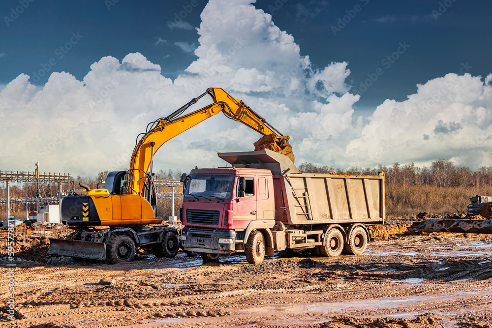 A wheeled excavator loads a dump truck with soil and sand. An excavator with a high-raised bucket against a cloudy sky View from the trench. Removal of soil from a construction site or quarry. A wheeled excavator loads a dump truck with soil and sand. An excavator with a high-raised bucket against a cloudy sky View from the trench. Removal of soil from a construction site or quarry.