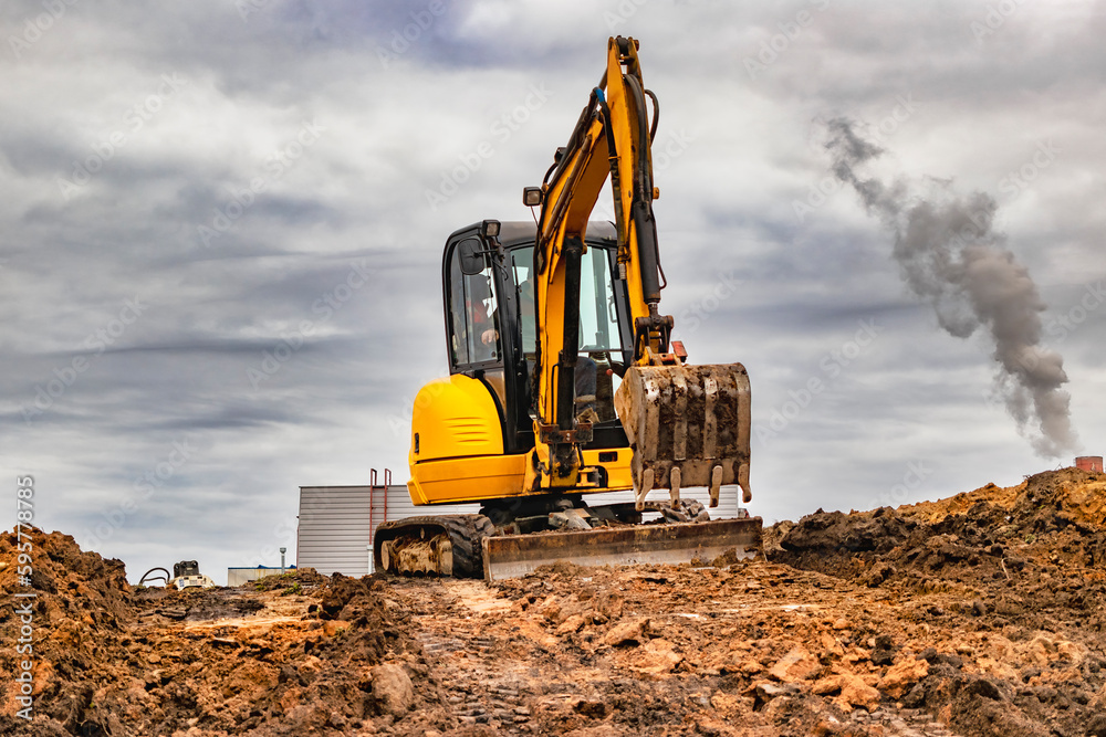 Mini excavator at the construction site on the edge of a pit against a cloudy blue sky. Compact construction equipment for earthworks. An indispensable assistant for earthworks. Mini excavator at the construction site on the edge of a pit against a cloudy blue sky. Compact construction equipment for earthworks. An indispensable assistant for earthworks.