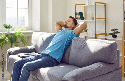Man relaxing on a comfortable couch at home. Young guy in a blue T shirt sitting on a gray couch with his hands behind his head and a laptop beside him. Relaxation, break, pause, free time concept