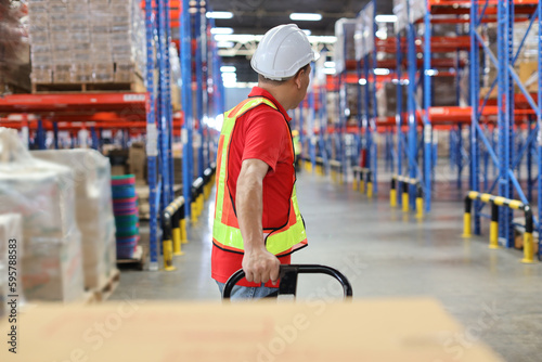 Rear view warehouse workers man with hardhats and reflective jackets pulling a pallet truck and taking or upload with large box package to shelf in retail warehouse logistics, distribution center
