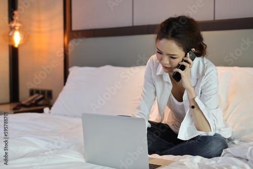 Active and unhappy asian woman in casual sitting on bed and working smart mobile phone and computer in hotel, beauty businesswoman with bedroom background. Lifestyle technology concept