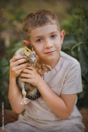The boy tenderly holds a duck in his hands. A lovely warm summer evening. Sunny day. The kid presses a little duckling to his cheek. Fairy tale. The kid sits near a fabulous tree and holds a duck in h