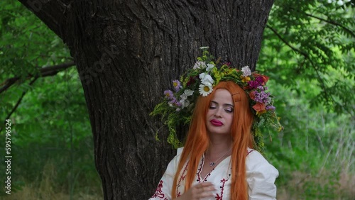 Beautiful girl with long red hair with flower crown in white and red embroidered ethnic suit stands by tree. Fairytale character, forest spirit, mavka, fairy. 4K 