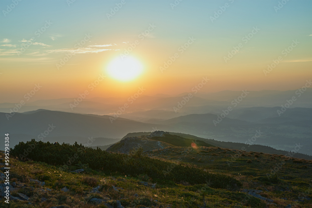 Obraz premium Sunrise in mountains. Natural mountain landscape with illuminated misty peaks, foggy slopes and valleys, blue sky with orange yellow sunlight