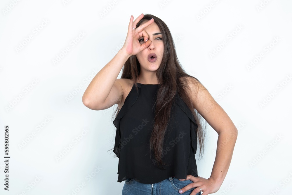 young brunette woman wearing black blouse over white studio background doing ok gesture shocked with surprised face, eye looking through fingers. Unbelieving expression. young brunette woman wearing black blouse over white studio background doing ok gesture shocked with surprised face, eye looking through fingers. Unbelieving expression.