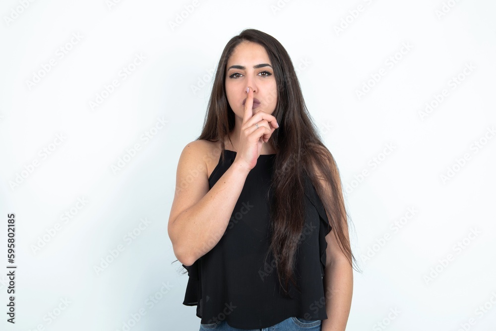 young brunette woman wearing black blouse over white studio background makes silence gesture, keeps finger over lips. Silence and secret concept. young brunette woman wearing black blouse over white studio background makes silence gesture, keeps finger over lips. Silence and secret concept.