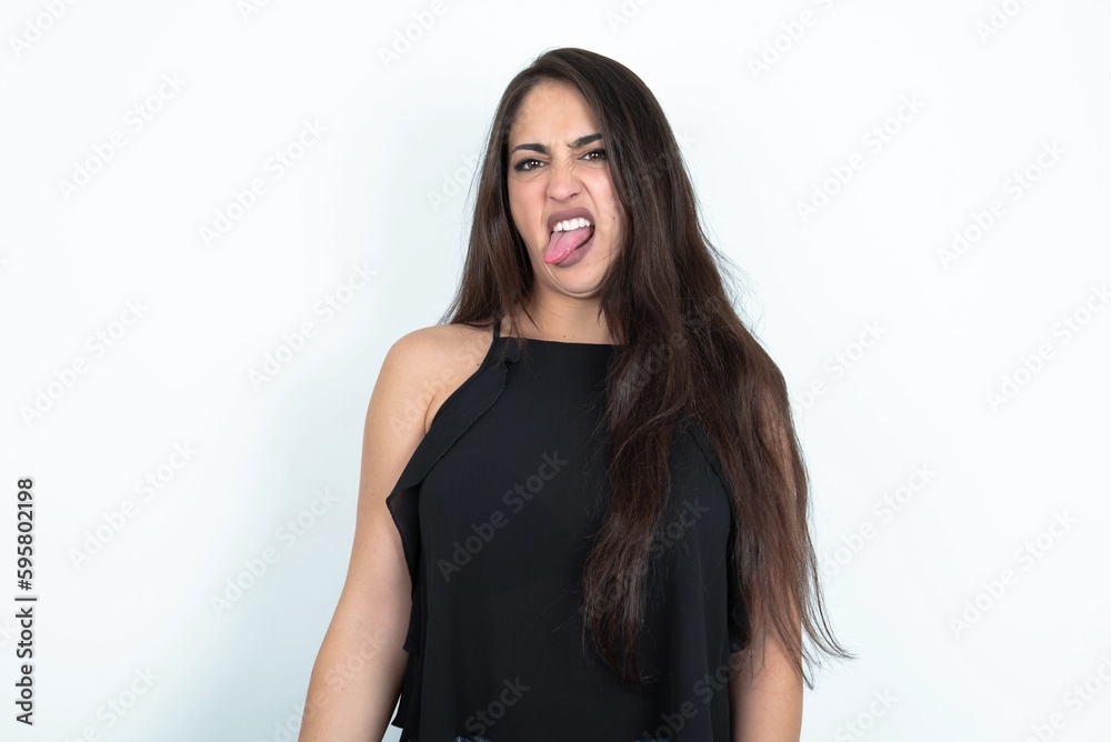 Naklejka premium young brunette woman wearing black blouse over white studio background sticking tongue out happy with funny expression. Emotion concept.