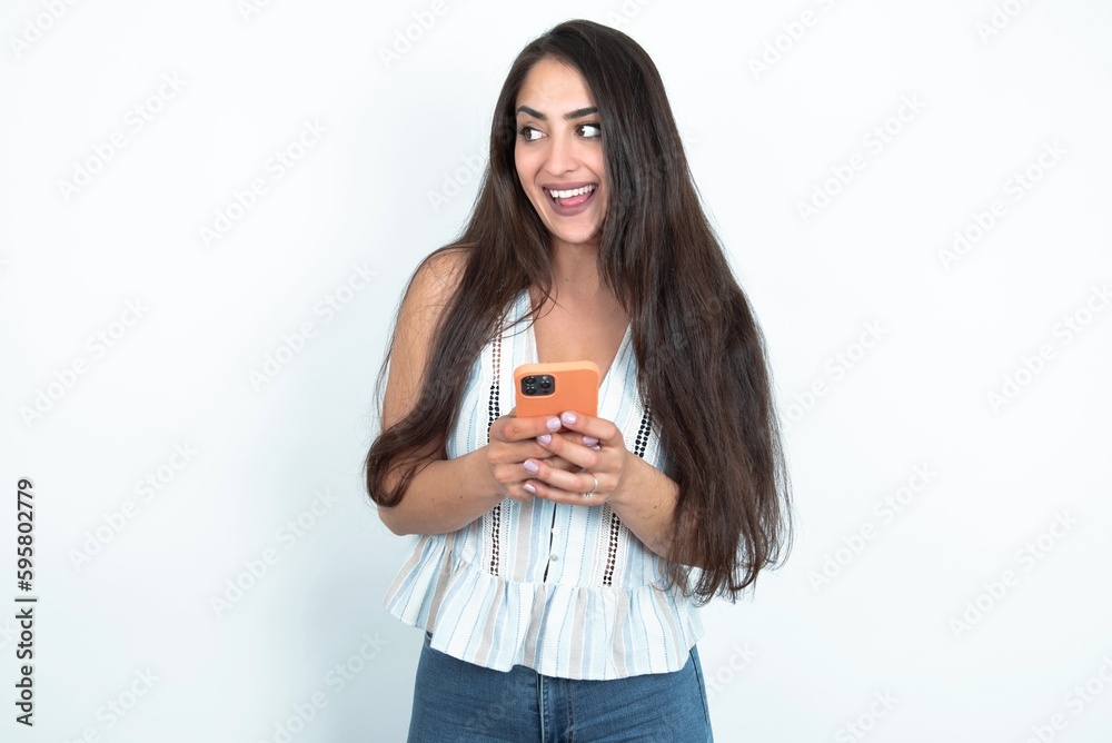 young brunette woman wearing white tank top over white studio background holding a smartphone and looking sideways at blank copyspace. young brunette woman wearing white tank top over white studio background holding a smartphone and looking sideways at blank copyspace.