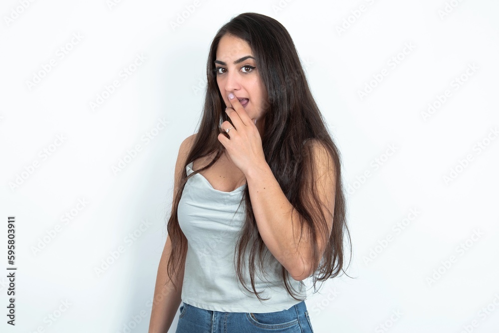 young brunette woman wearing white tank top over white studio background covers mouth and looks with wonder at camera, cannot believe unexpected rumors. young brunette woman wearing white tank top over white studio background covers mouth and looks with wonder at camera, cannot believe unexpected rumors.