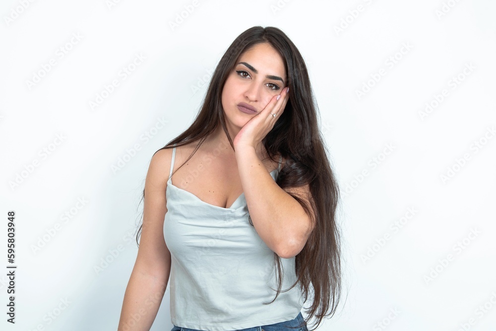 Sad lonely young brunette woman wearing white tank top over white studio background touches cheek with hand bites lower lip and gazes with displeasure. Bad emotions Sad lonely young brunette woman wearing white tank top over white studio background touches cheek with hand bites lower lip and gazes with displeasure. Bad emotions