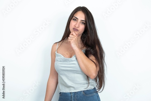 young brunette woman wearing white tank top over white studio background looking confident at the camera smiling with crossed arms and hand raised on chin. Thinking positive. © Jihan