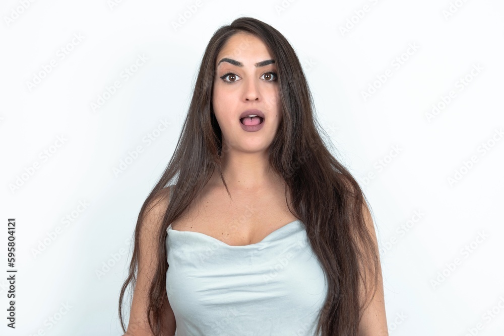 Shocked young brunette woman wearing white tank top over white studio background stares bugged eyes keeps mouth opened has surprised expression. Omg concept