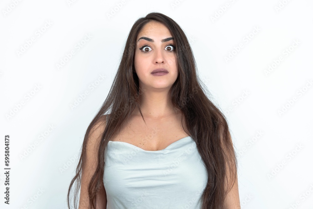 Stunned brunette woman wearing white tank top over white studio background stares reacts on shocking news. Astonished girl holds breath Stunned brunette woman wearing white tank top over white studio background stares reacts on shocking news. Astonished girl holds breath