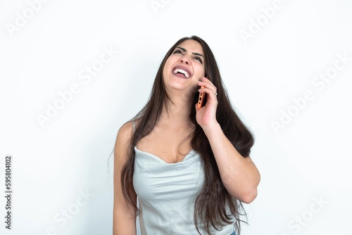 Funny young brunette woman wearing white tank top over white studio background laughs happily, has phone conversation, being amused by friend, closes eyes.
