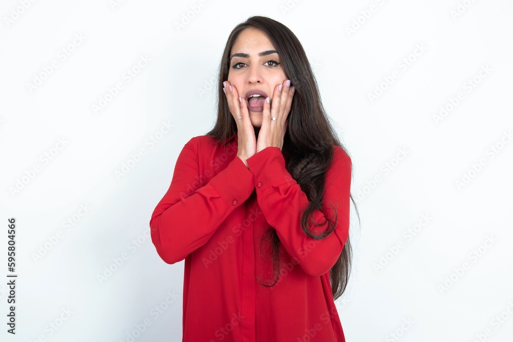 Upset Young brunette woman wearing red shirt over white studio background touching face with two hands Upset Young brunette woman wearing red shirt over white studio background touching face with two hands
