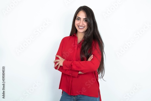 Portrait of charming Young brunette woman wearing red shirt over white studio background standing confidently smiling toothily with hands folded