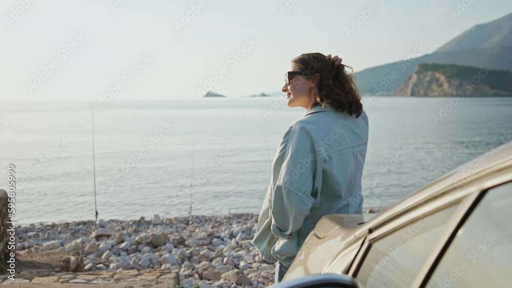 A young Caucasian woman leaning against her car parked by the sea and enjoying the beautiful view. A young Caucasian woman leaning against her car parked by the sea and enjoying the beautiful view.