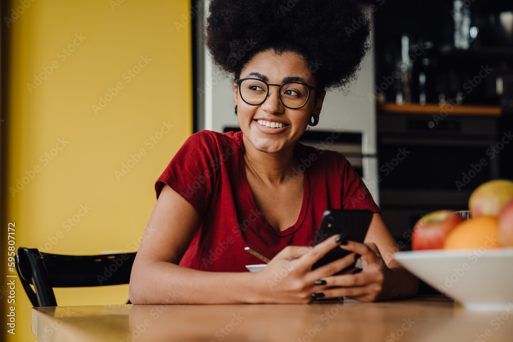 African woman using smartphone while having breakfast in kitchen