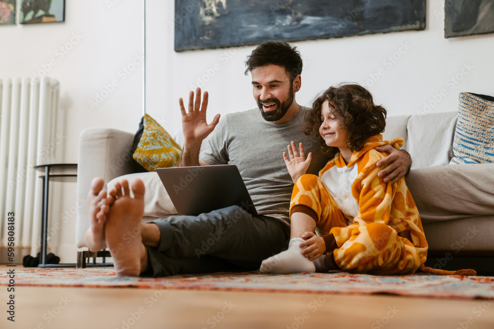 Father and son waving hands while making video call via laptop sitting on floor in living room Father and son waving hands while making video call via laptop sitting on floor in living room