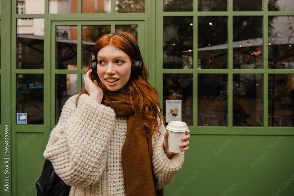 Fototapeta premium Smiling redhead woman listening music with headphones and drinking coffee while standing outdoors