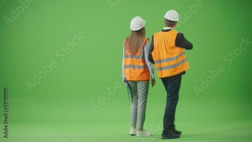 Green Screen.Female and Male Workers Partners in Safety Jackets and White Helmets,Holding Tablet and Paper Clipboard Walking Through Discussing Project Standing Back to Camera.Team Work Concept.